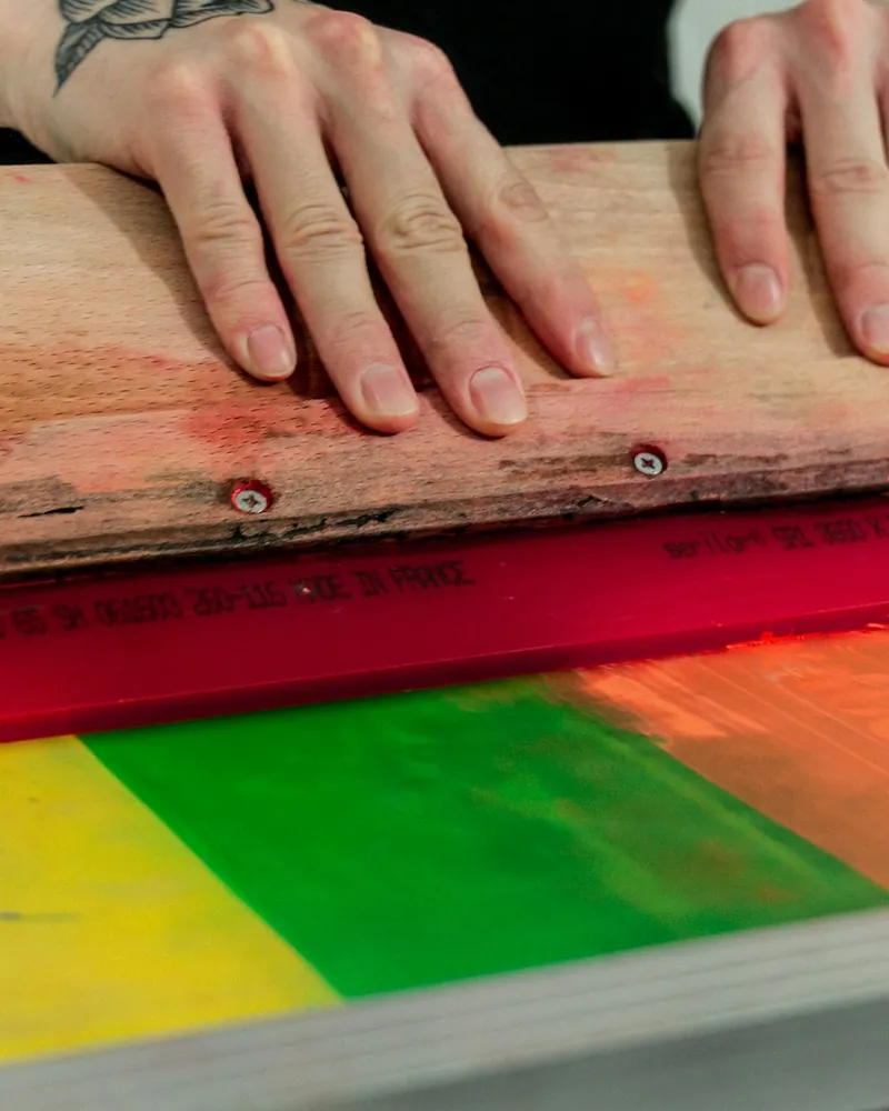 Racleta de goma roja apoyada sobre una pantalla de serigrafía con franjas de tinta amarilla, verde y naranja