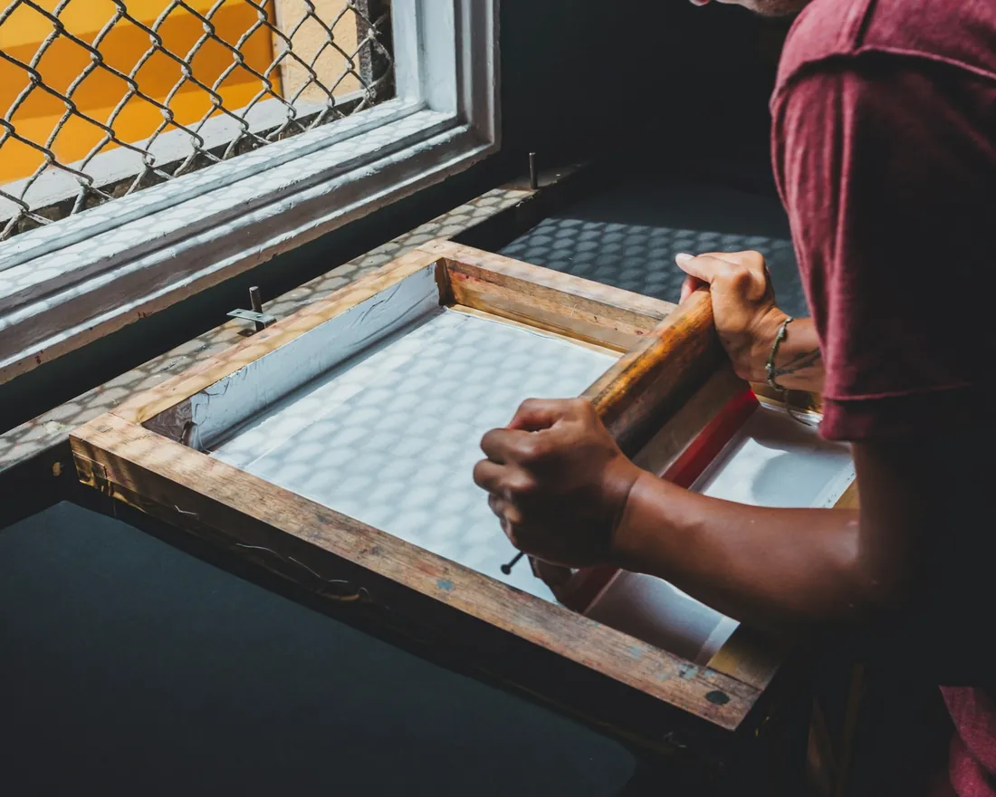 Bastidor de madera con malla de serigrafía apoyado junto a una ventana mientras unas manos sujetan una racleta de mango de madera